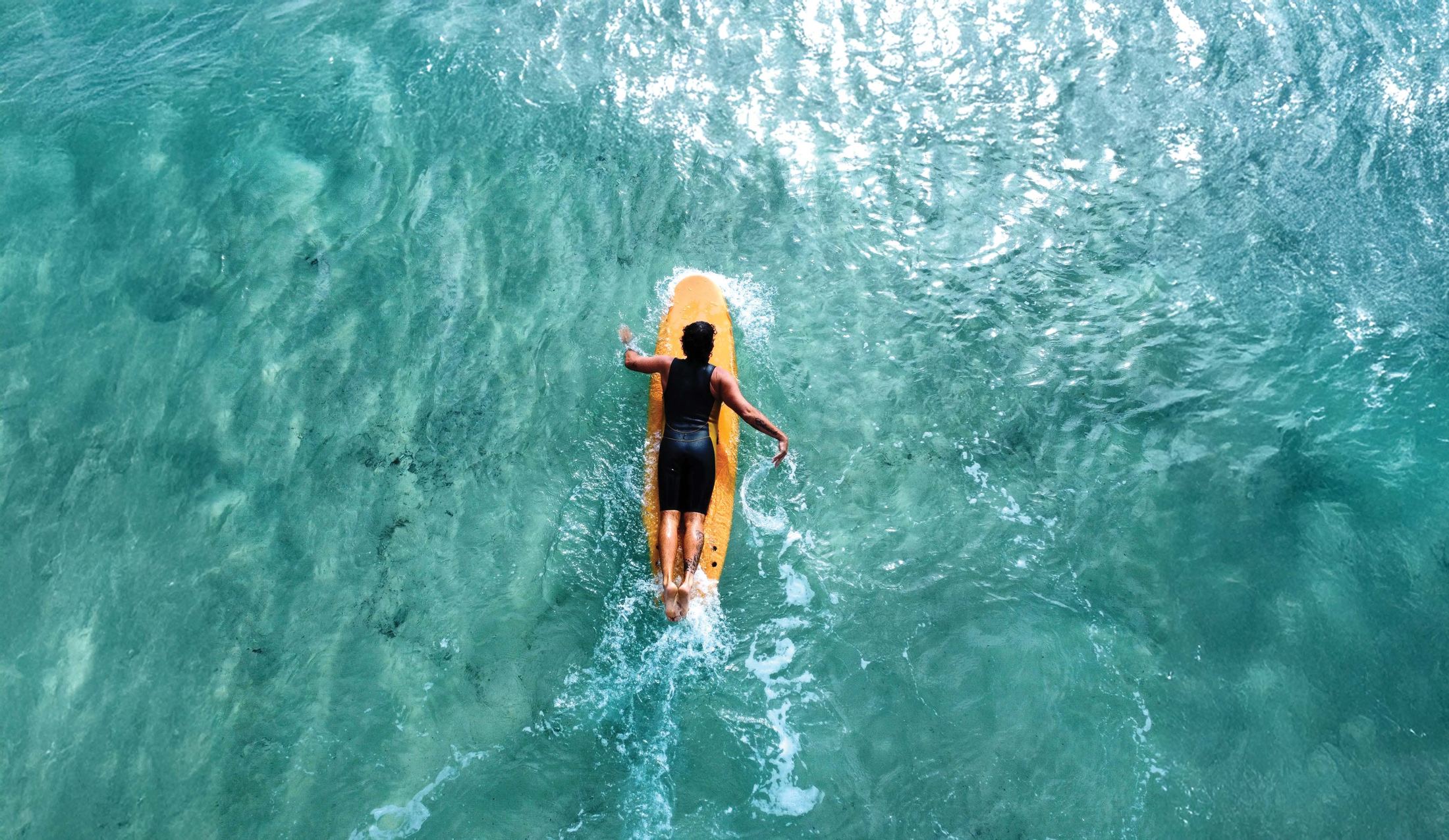 Surfer paddling a softboard over clear turquoise water, seen from above